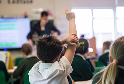 Child with his hand up to answer a question at the back of the classroom